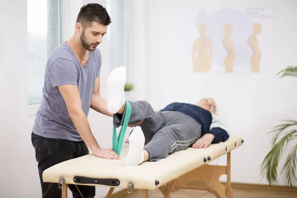 Hombre de edad avanzada gris se encuentra en una mesa de fisioterapia, y el joven médico le ayuda durante los ejercicios —  Fotos de Stock