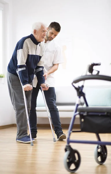 Älterer Mann geht auf Krücken und wird von einer hilfsbereiten Krankenschwester unterstützt — Stockfoto