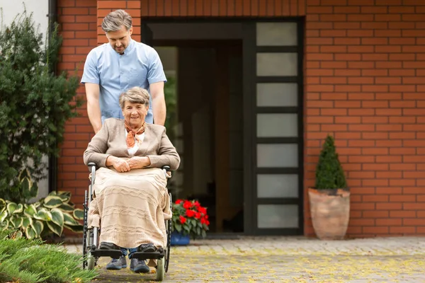 Sorridente donna anziana sulla sedia a rotelle sostenuta da caregiver di fronte alla casa — Foto Stock