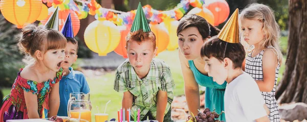 Vedere panoramică a băiețelului roșcat, animator de petrecere și elevi suflând lumânări pe tort de ziua de naștere în timpul petrecerii de grădină pentru copii — Fotografie, imagine de stoc
