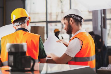 Teslim belgelerini inceleyen turuncu güvenlik ceketve kasklı iki fabrika işçisi