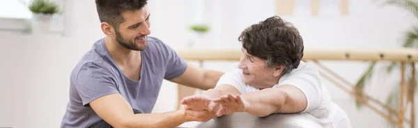 Panoramatická fotka mladého fyzioterapeuta pomáhající staré ženě při cvičení — Stock fotografie
