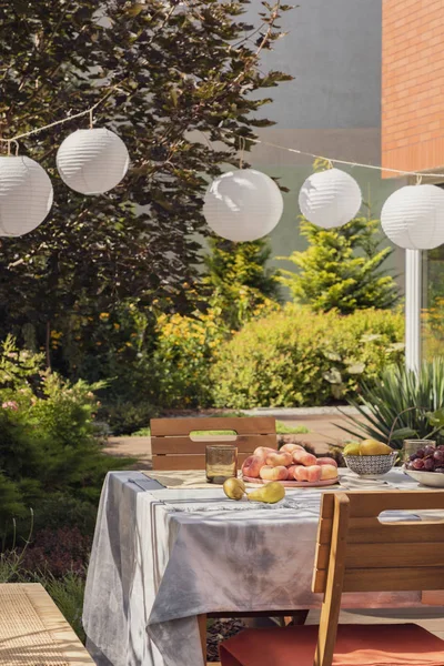 Trästolar vid bordet med frukt i trädgården med växter och vita lyktor under festen. Verkligt foto — Stockfoto