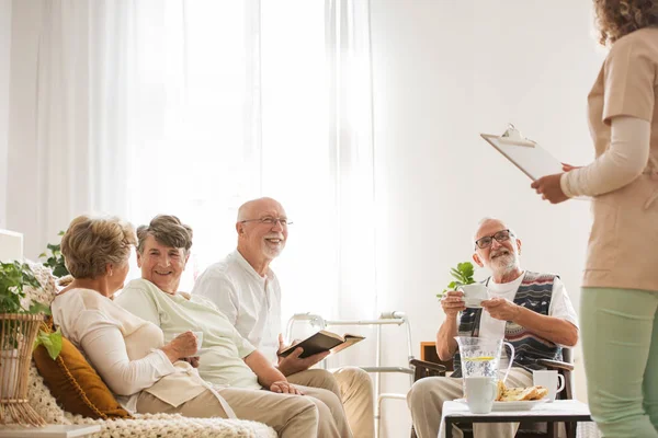 Grupp av äldre sjukhem pensionärer sitter tillsammans på gemensamt vardagsrum lyssnar på ungSjuksköterska — Stockfoto