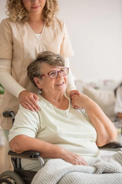 Dame aînée en fauteuil roulant avec une jeune bénévole en uniforme beige qui la soutient — Photo