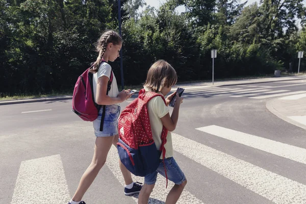 Schoolboy with backpack and his teenage sister using mobile phones on pedestrian crossing — Stock Photo, Image