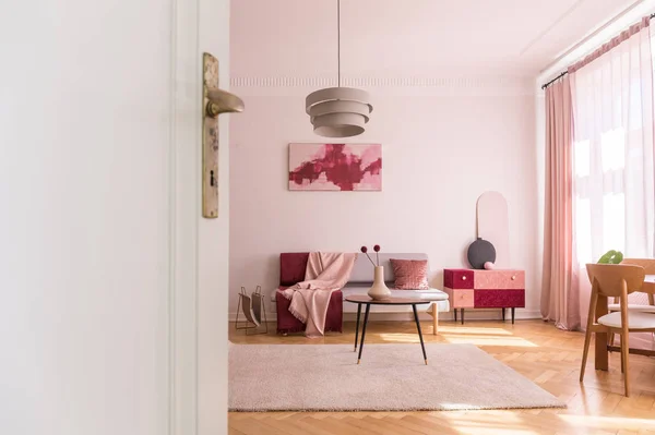 Białe drzwi salonu z plakatem nad kanapą w pobliżu stołu z kwiatami na różowym dywanie. Prawdziwe zdjęcie — Zdjęcie stockowe