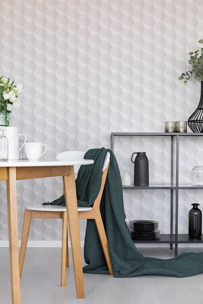 Dark green blanket on wooden chair next to table in stylish dining room interior with metal shelf with plates and jugs and geometric pattern on wallpaper