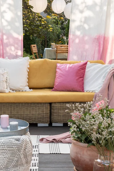 Flores y velas frente al sofá de ratán con almohadas de colores en la terraza con cortinas. Foto real —  Fotos de Stock