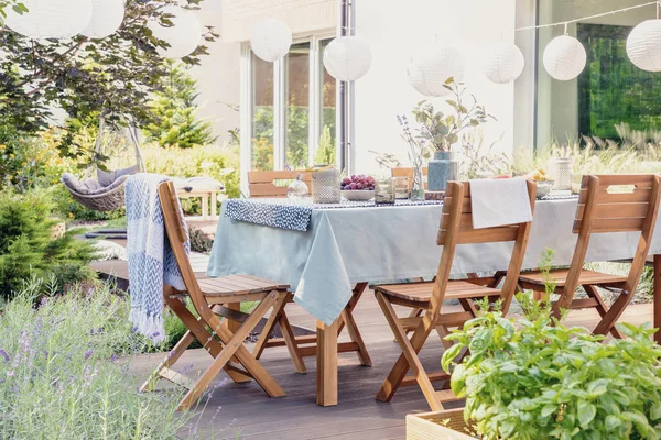Houten stoelen aan tafel met bloemen en eten in de tuin met planten en lantaarns. Echte foto — Stockfoto