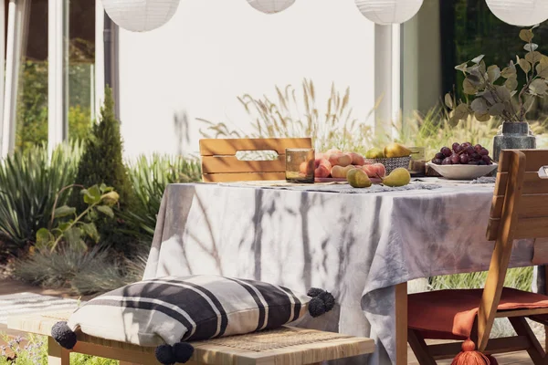 Mönstrad kudde på träbänk vid bord med frukt på husets terrass med växter. Riktiga foto — Stockfoto