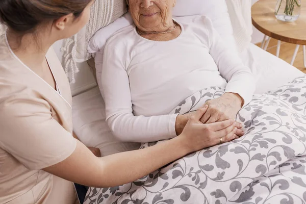 Mujer mayor positiva acostada en la cama, médico servicial en uniforme beige que la apoya —  Fotos de Stock