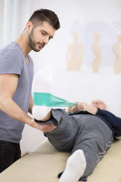 Homme âgé gris repose sur une table de physiothérapie, et le jeune médecin l'aide pendant les exercices — Photo