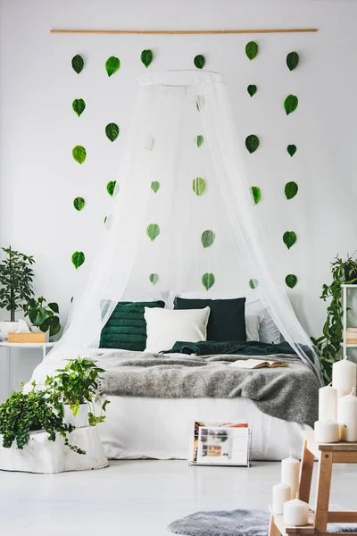 Witte slaapkamer interieur met kingsize hemelbed met smaragdgroene kussens en grijze deken, stedelijke jungle en groene bladeren aan de muur — Stockfoto