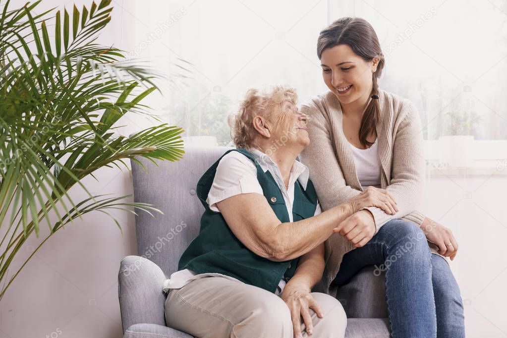 Senior lady sitting in armchair at nursing home, supporting nurse behind her