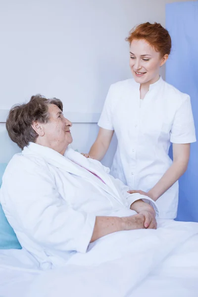 Ältere graue Frau liegt im weißen Krankenhausbett mit junger hilfsbereiter Krankenschwester, die ihre Hand hält — Stockfoto
