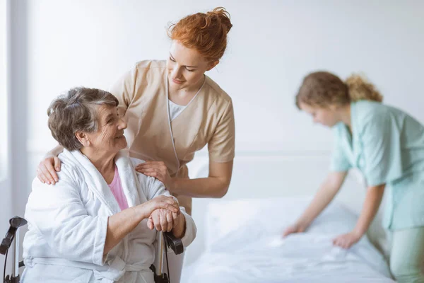 Oudere vrouw op rolstoel in verpleeghuis met behulpzame arts aan haar kant en jonge verpleegster het maken van het bed — Stockfoto