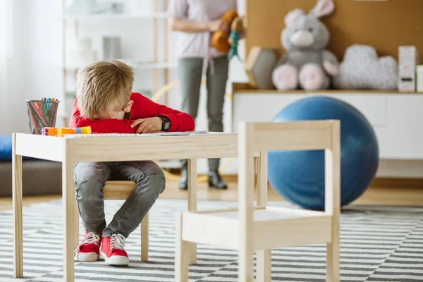 Lindo niño vistiendo suéter rojo sentado en una pequeña mesa de madera — Foto de Stock