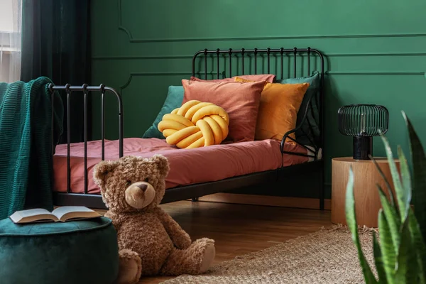 子供の寝室のインテリアの金属フレームベッドの前に緑のベルベットのプーフの大きな、茶色のテディベアとオープンブック。実際の写真 — ストック写真