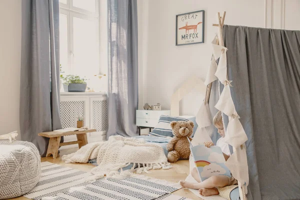 Véritable photo d'un intérieur bleu de chambre d'enfant avec matelas, rideaux, ours en peluche et tente où un garçon lit un livre — Photo