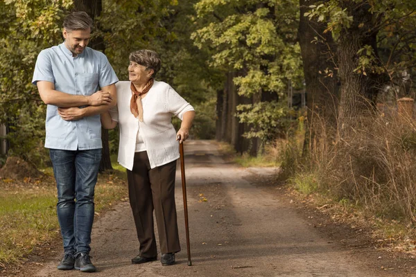 Enfermera masculina apoyando a anciana feliz con bastón en el bosque — Foto de Stock