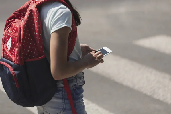 Primer plano en el niño con la mochila usando el teléfono inteligente mientras camina a través del paso de peatones —  Fotos de Stock
