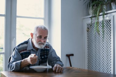 Gri saçlı ve sakallı yaşlı Büyükbaba mutfakta yalnız oturuyor kahvaltı yiyor