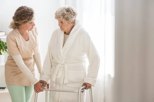 Mujer mayor en albornoz con andador y enfermera servicial apoyándola —  Fotos de Stock