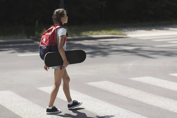 Девушка с рюкзаком и скейтбордом идет через пешеходный переход в школу — стоковое фото