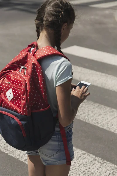 Niño con mochila usando teléfono inteligente mientras camina a través del cruce peatonal — Foto de Stock