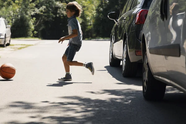 Niño descuidado corriendo detrás de la pelota en la carretera junto a los coches —  Fotos de Stock