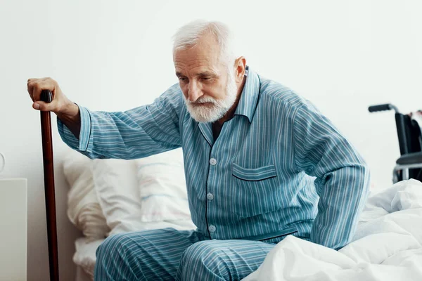 Viejo enfermo con barba gris y pelo con pijama azul y sentado en la cama en casa —  Fotos de Stock