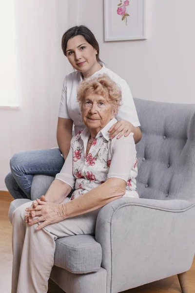 Старшая леди, сидящая в кресле в доме престарелых, поддерживающая медсестру позади нее — стоковое фото