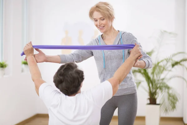 Fisioterapeuta sonriente ayudando a las mujeres mayores a ejercitarse con bandas de resistencia — Foto de Stock