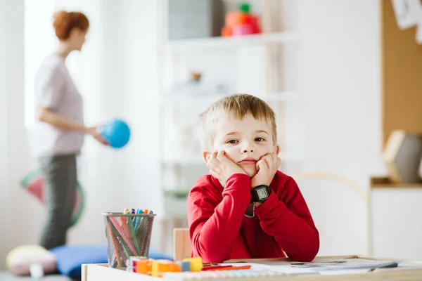 Söt liten pojke klädd i röd tröja sitter vid små träbord — Stockfoto