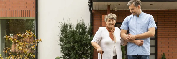 Вид счастливой пожилой дамы с тростью, идущей на прогулку с красивым мужчиной-медсестрой в саду дома престарелых — стоковое фото