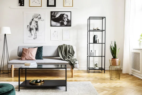 Mesa frente al sofá junto a la lámpara en el interior del apartamento blanco con planta y carteles. Foto real — Foto de Stock