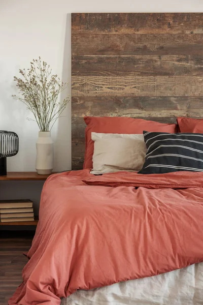 Bloemen in beige vaas en zwarte industriële lamp op houten nachtkastje naast comfortabel bed met kussens — Stockfoto