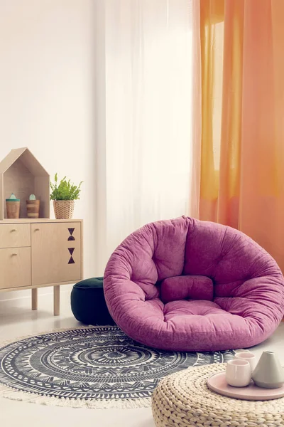 Pink cozy pouf in the middle of fashionable natural kid\'s room with wooden furniture and ethno carpet on white floor