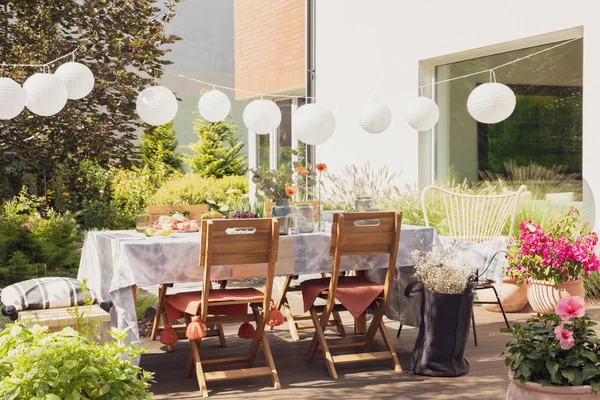 Fehér lámpák asztal felett és fa székek a teraszon, virágokkal a ház mellett — Stock Fotó