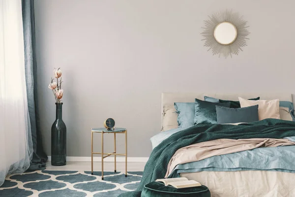 Flowers in stylish bottle like vase next to trendy nightstand with clock in beautiful bedroom interior with beige and emerald green bedding — Stock Image Flowers in stylish bottle like vase next to trendy nightstand with clock in beautiful bedroom interior with beige and emerald green bedding Stock Photo