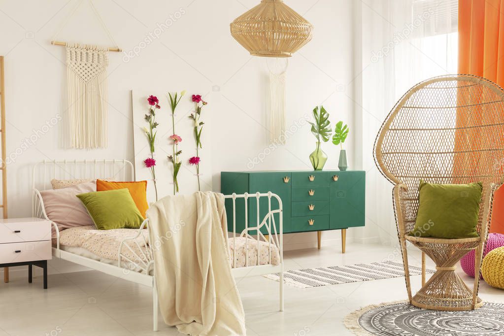 Peacock chair with olive green pillow in trendsetting bedroom interior with white metal bed with colorful pillows and handmade flower board and macrame