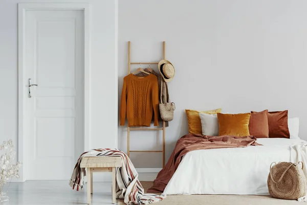 在特大号床旁边，在木床上戴毛衣、柳条帽和包，白色卧室内部配有天鹅绒枕头，空墙上有复制空间 — 图库照片