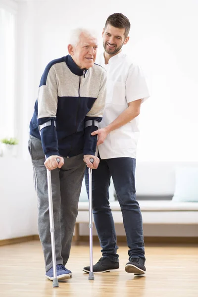 Uomo anziano che cammina con le stampelle e un infermiere di sesso maschile che lo sostiene — Foto Stock