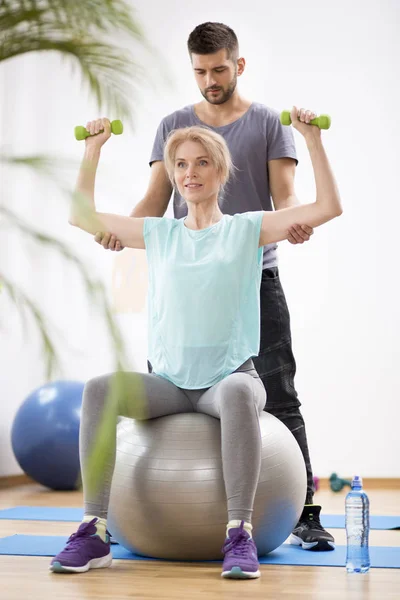 Mujer rubia de mediana edad haciendo ejercicio en la pelota de gimnasia durante la sesión con el fisioterapeuta —  Fotos de Stock