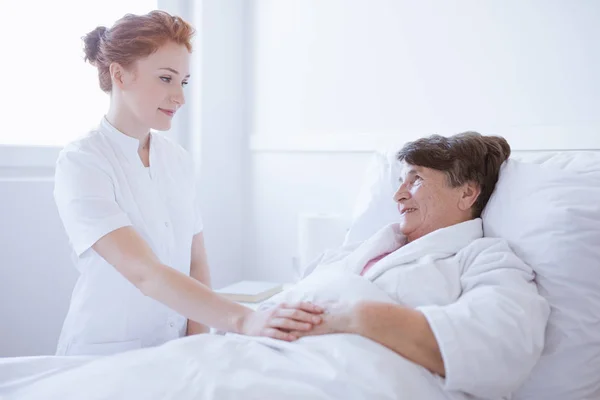 Ανώτερη γκρίζα γυναίκα που κείτεται σε λευκό νοσοκομείο κρεβάτι με νεαρή χρήσιμη νοσοκόμα κρατώντας το χέρι της — Φωτογραφία Αρχείου