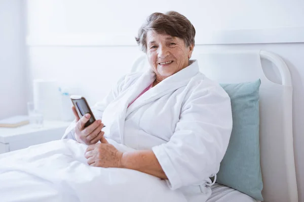 Sonriendo señora mayor sosteniendo el teléfono móvil mientras está sentada en la cama del hospital — Foto de Stock
