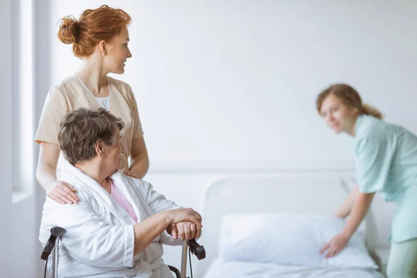 Ηλικιωμένη γυναίκα σε αναπηρικό καροτσάκι στο γηροκομείο με εξυπηρετικό γιατρό στο πλευρό της και νεαρή νοσοκόμα που φτιάχνει το κρεβάτι — Φωτογραφία Αρχείου