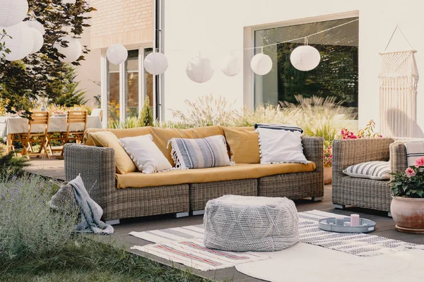 Puf junto a sofá de ratán y sillón en terraza de madera con flores y lámparas — Foto de Stock