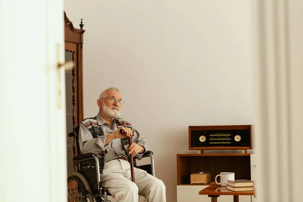 Starší muž s šedými vlasy a vousy a s holí sedící na lenochodě v bytě — Stock fotografie